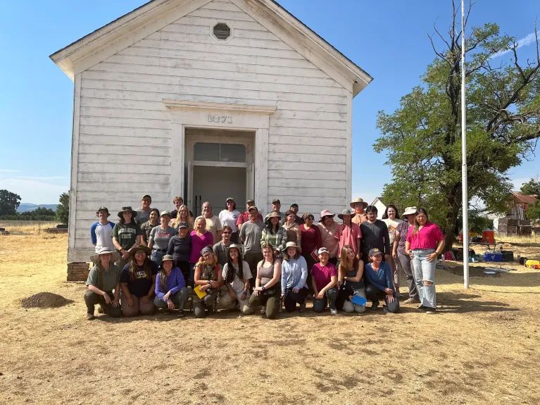 Summer 2024 students standing in front of a white single room schoolhouse